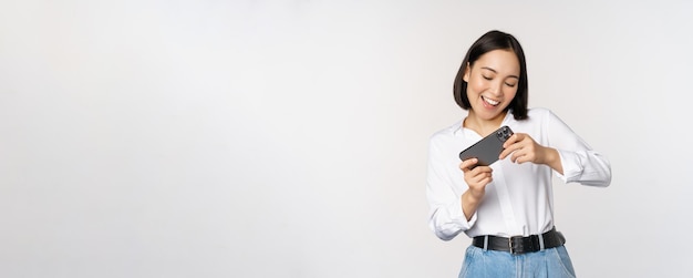 Joven mujer coreana asiática jugando videojuegos móviles en un teléfono inteligente mirando la pantalla horizontal del teléfono de pie sobre fondo blanco