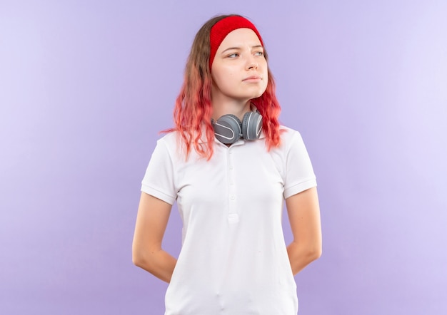 Joven mujer deportiva con auriculares mirando a un lado con expresión de confianza de pie sobre la pared púrpura