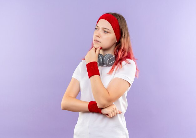 Joven mujer deportiva con auriculares mirando a un lado con la mano en la barbilla pensando de pie sobre la pared púrpura