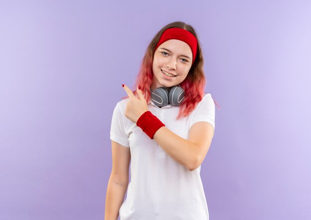 Joven mujer deportiva con auriculares sonriendo alegremente apuntando hacia atrás de pie sobre la pared púrpura