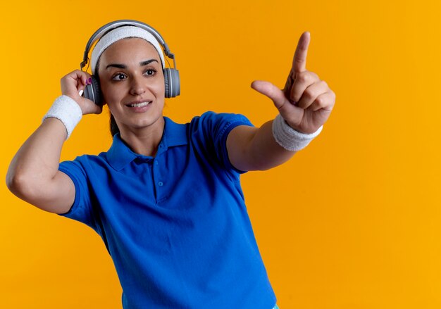 Joven mujer deportiva caucásica complacida con diadema y muñequeras en auriculares apunta hacia adelante aislado sobre fondo naranja con espacio de copia