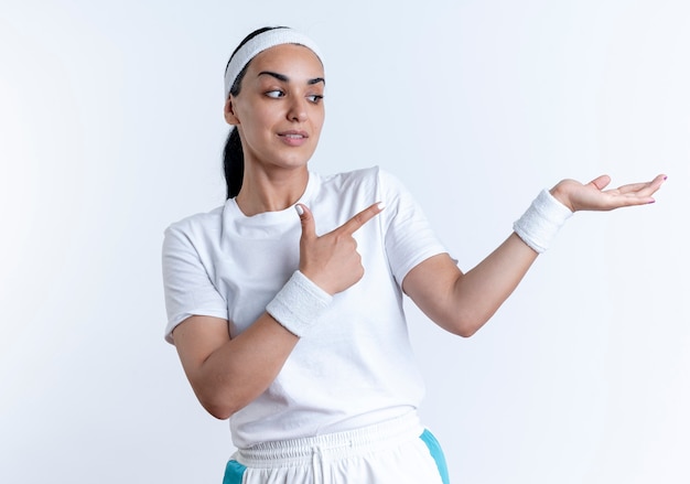 Joven mujer deportiva caucásica confiada con diadema y muñequeras puntos en la mano vacía aislada en el espacio en blanco con espacio de copia