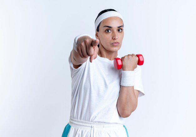 Joven mujer deportiva caucásica confiada con diadema y muñequeras sostiene pesas y apunta a la cámara aislada en el espacio en blanco con espacio de copia