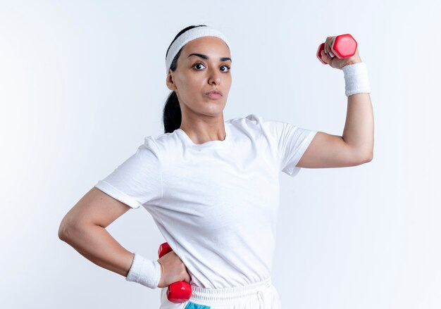 Joven mujer deportiva caucásica confiada con diadema y muñequeras tensa bíceps sosteniendo pesas aislado en espacio en blanco con espacio de copia