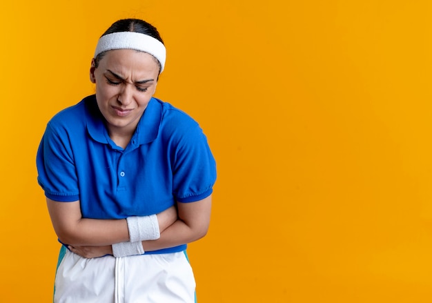 Joven mujer deportiva caucásica dolorida con diadema y muñequeras sostiene el vientre en naranja con espacio de copia