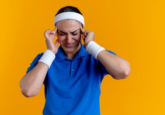 Joven mujer deportiva caucásica molesta con diadema y muñequeras sostiene la cabeza con los ojos cerrados en naranja con espacio de copia