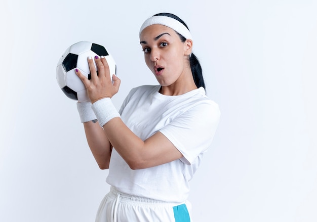 Joven mujer deportiva caucásica sorprendida con diadema y muñequeras sostiene la bola aislada en el espacio en blanco con espacio de copia