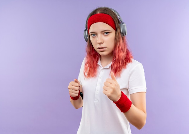 Joven mujer deportiva en diadema con auriculares con cara seria, entrenando con brazalete de smartphone parado sobre pared púrpura
