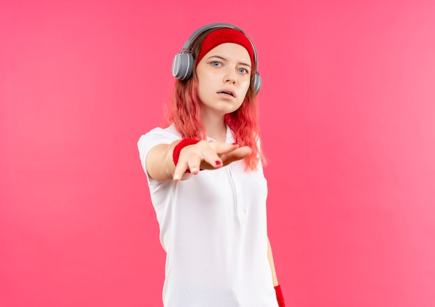 Foto gratuita joven mujer deportiva en diadema con auriculares preocupado gesticulando con la mano sobre la pared rosa