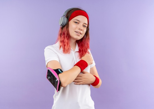 Joven mujer deportiva en diadema con auriculares sonriendo con brazalete de smartphone parado sobre pared púrpura