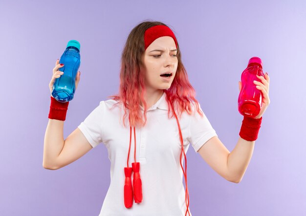 Joven mujer deportiva en diadema con cuerda alrededor de su cuello sosteniendo dos botellas de agua con aspecto confundido tratando de tomar una decisión de pie sobre la pared púrpura