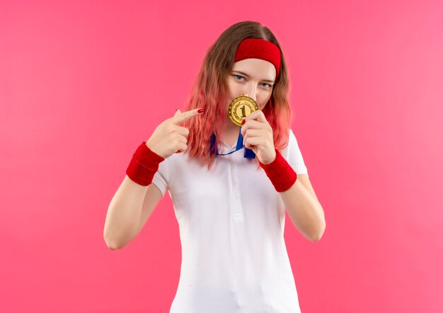 Joven mujer deportiva en diadema mostrando medalla de oro apuntando con el dedo hacia ella mirando confiado de pie sobre la pared rosa