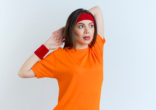 Joven mujer deportiva con diadema y muñequeras manteniendo las manos juntas detrás de la cabeza mirando al lado ejercicio aislado en la pared blanca con espacio de copia