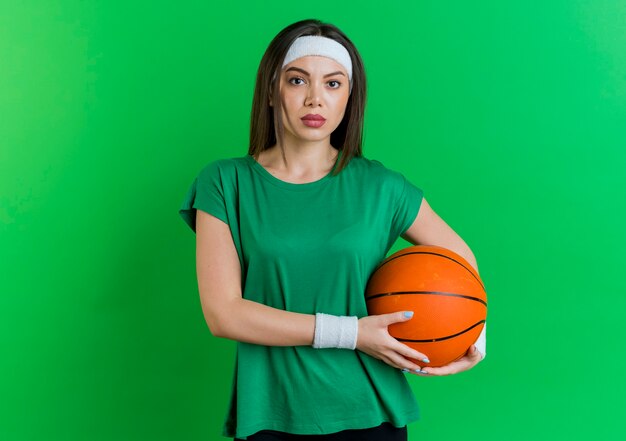 Joven mujer deportiva con diadema y muñequeras sosteniendo una pelota de baloncesto y mirando