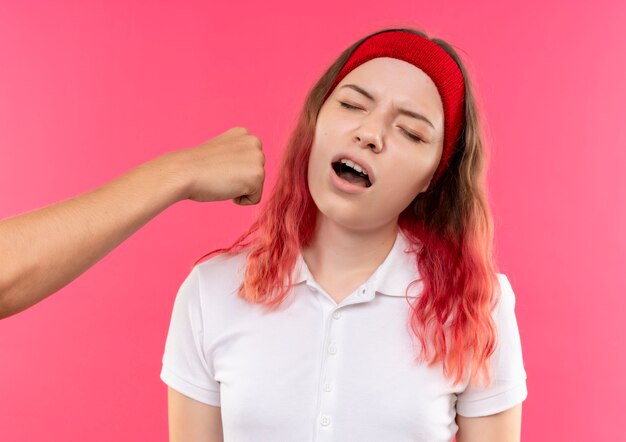 Joven mujer deportiva en diadema recibiendo puch en la cara con puño parado sobre pared rosa