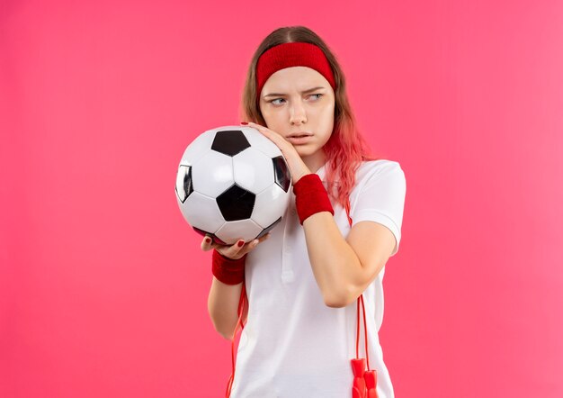 Joven mujer deportiva en diadema sosteniendo un balón de fútbol mirando a un lado con expresión de miedo de pie sobre la pared rosa