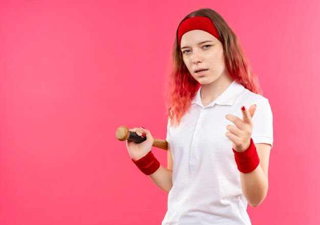 Joven mujer deportiva en diadema sosteniendo un bate de béisbol mirando confiado apuntando con el dedo a la cámara de pie sobre la pared rosa