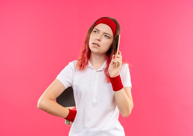 Joven mujer deportiva en diadema sosteniendo dos raquetas de tenis de mesa mirando a un lado con expresión pensativa en la cara de pie sobre la pared rosa