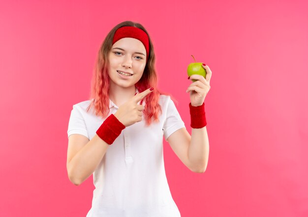 Joven mujer deportiva en diadema sosteniendo manzana verde apuntando con el dedo a ella sonriendo confiados de pie sobre la pared rosa