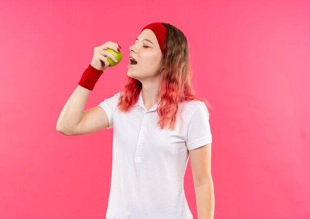 Joven mujer deportiva en diadema sosteniendo manzana verde va a morderla de pie sobre la pared rosa