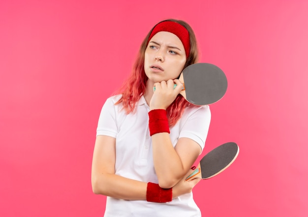 Joven mujer deportiva en diadema sosteniendo la raqueta de tenis de mesa mirando a un lado con expresión pensativa en la cara pensando de pie sobre la pared rosa