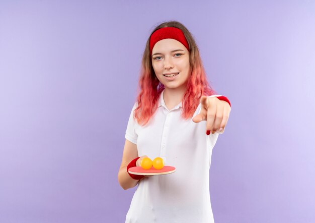 Joven mujer deportiva en diadema sosteniendo raqueta de tenis de mesa y pelotas apuntando con el dedo a la cámara sonriendo de pie sobre la pared púrpura