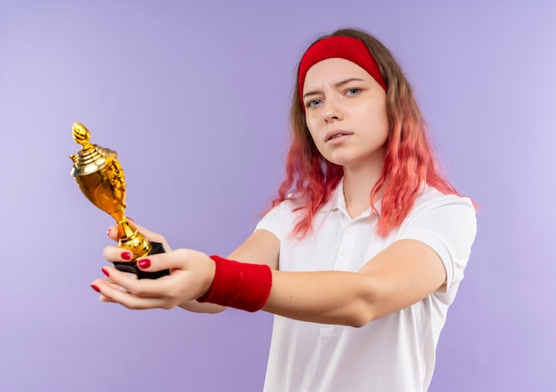 Foto gratuita joven mujer deportiva en diadema sosteniendo trofeo con expresión seria de pie sobre la pared púrpura