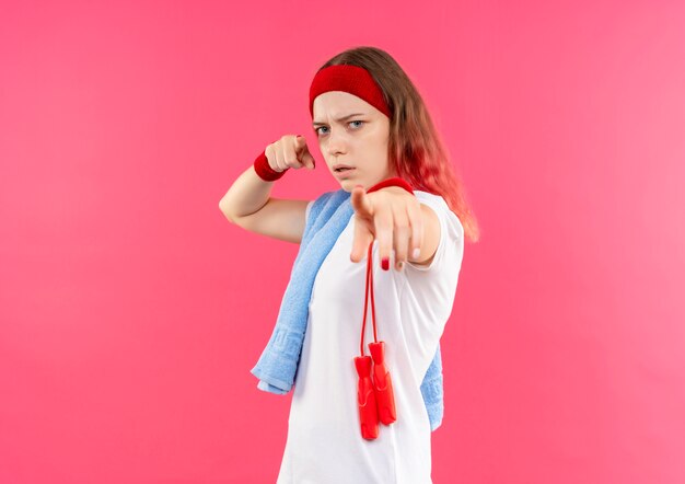 Joven mujer deportiva en diadema con toalla en el hombro apuntando con el dedo a la cámara con cara seria de pie sobre la pared rosa