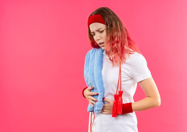 Joven mujer deportiva en diadema con toalla en el hombro con aspecto cansado y agotado tocando su vientre parado sobre pared rosa