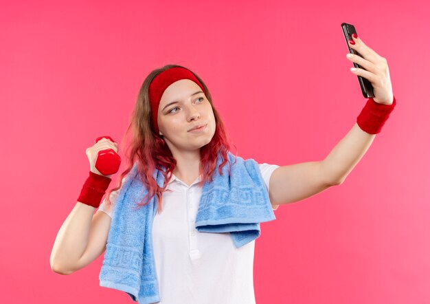 Joven mujer deportiva en diadema con toalla en el hombro tomando un selfie de sí misma mostrando mancuernas en la mano a la cámara de su teléfono inteligente de pie sobre la pared rosa