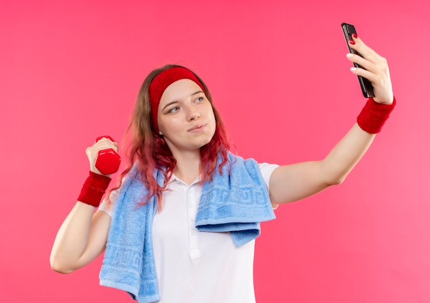 Joven mujer deportiva en diadema con toalla en el hombro tomando un selfie de sí misma mostrando mancuernas en la mano a la cámara de su teléfono inteligente de pie sobre la pared rosa