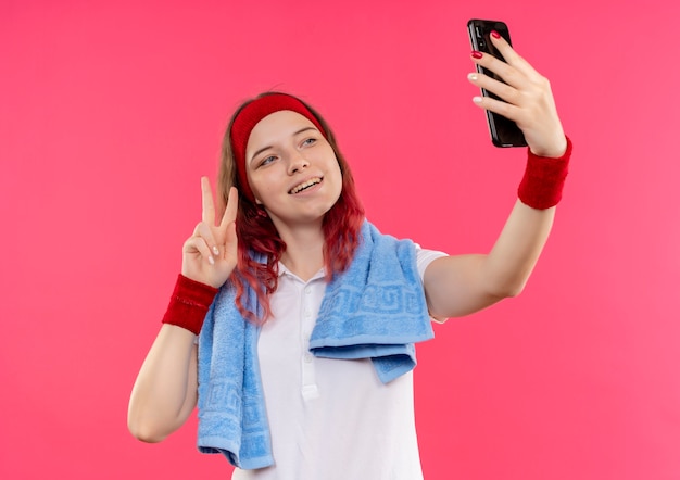 Foto gratuita joven mujer deportiva en diadema con toalla en el hombro tomando un selfie de sí misma mostrando el signo de la victoria a la cámara de su teléfono inteligente de pie sobre la pared rosa