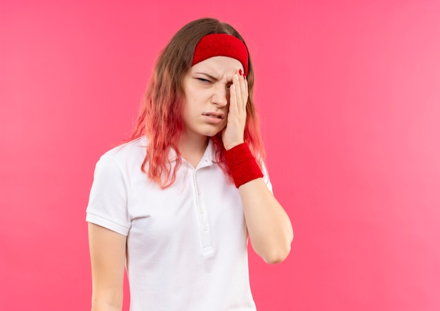 Joven mujer deportiva en diadema tocando su cabeza con aspecto cansado y aburrido que sufre de dolor de cabeza de pie sobre la pared rosa