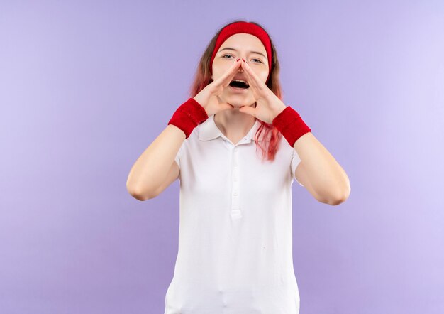 Joven mujer deportiva gritando o llamando a alguien con los brazos cerca de la boca de pie sobre la pared púrpura