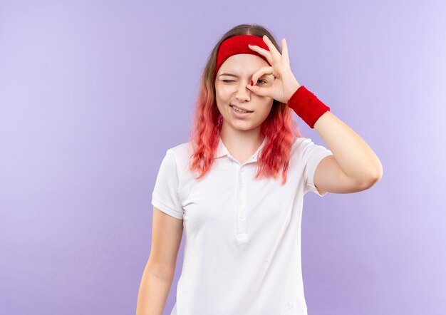 Joven mujer deportiva haciendo bien firmar con los dedos mirando a través de este cartel con una sonrisa en la cara de pie sobre la pared púrpura