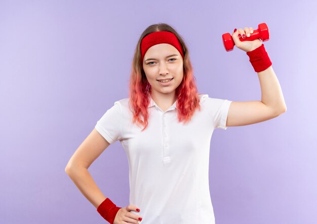 Joven mujer deportiva haciendo ejercicios con una mancuerna sonriendo confiados de pie sobre la pared púrpura