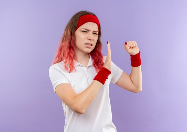 Joven mujer deportiva haciendo gesto de defensa con el brazo apretando el puño con expresión de disgusto de pie sobre la pared púrpura