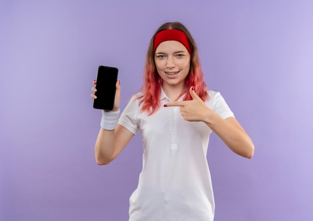 Joven mujer deportiva mostrando smartphone apuntando con el dedo con sonrisa de confianza de pie sobre la pared púrpura