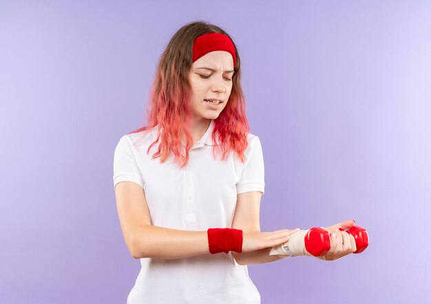 Joven mujer deportiva sosteniendo dos mancuernas haciendo ejercicios mirando su muñeca vendada sintiendo dolor de pie sobre la pared púrpura