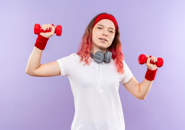 Joven mujer deportiva sosteniendo dos pesas haciendo ejercicios con sonrisa segura de pie sobre la pared púrpura