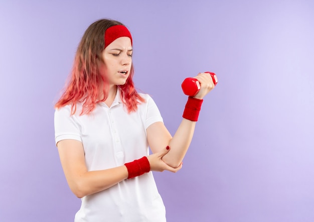 Joven mujer deportiva sosteniendo una mancuerna haciendo ejercicios mirando el codo sintiendo dolor de pie sobre la pared púrpura