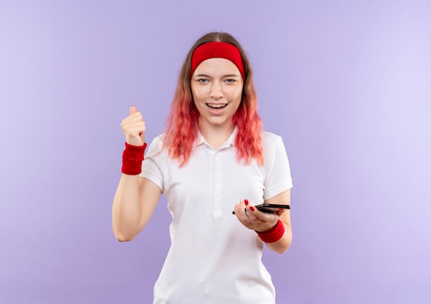 Joven mujer deportiva sosteniendo smartphone apretando el puño sonriendo alegremente de pie sobre la pared púrpura