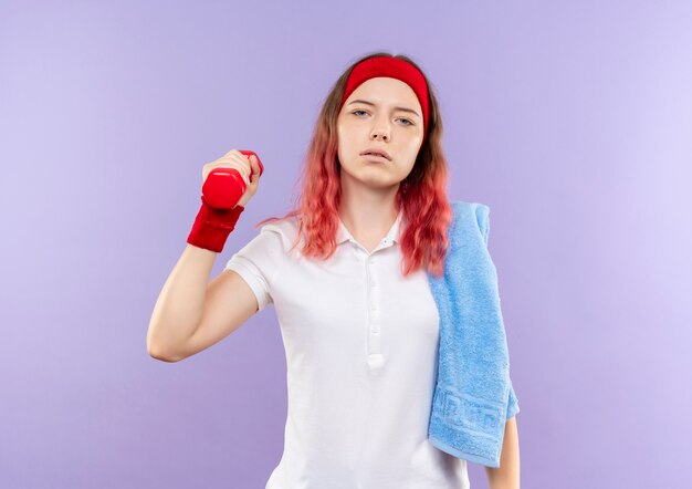 Joven mujer deportiva con una toalla en el hombro sosteniendo una pesa haciendo ejercicios con cara seria de pie sobre la pared púrpura
