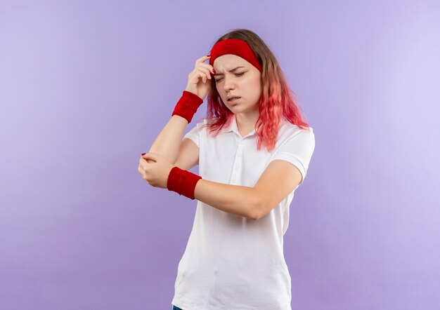 Joven mujer deportiva tocando su codo sintiendo dolor de pie sobre la pared púrpura