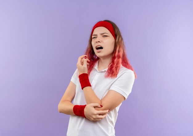 Joven mujer deportiva tocando su codo sintiendo dolor de pie sobre la pared púrpura