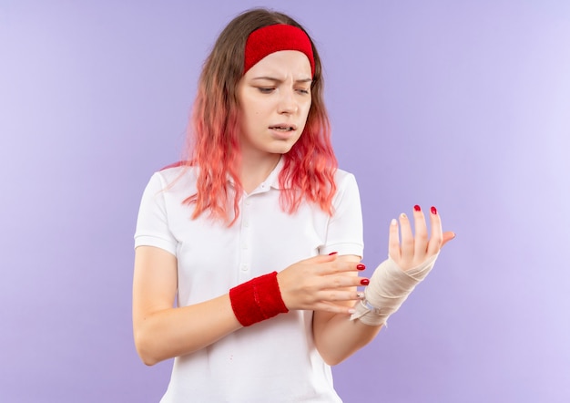 Joven mujer deportiva tocando su muñeca vendada sintiendo dolor de pie sobre la pared púrpura