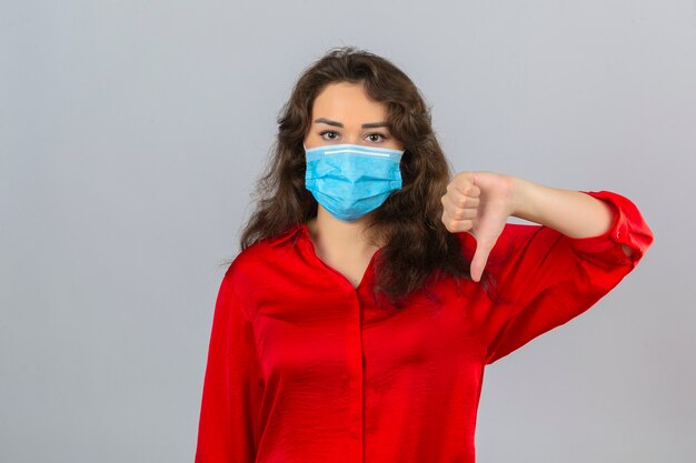 Joven mujer disgustada con blusa roja en máscara protectora médica mirando a la cámara mostrando el pulgar hacia abajo sobre fondo blanco aislado