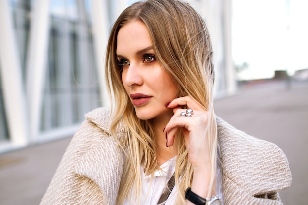 Joven mujer elegante bastante rubia posando en la calle, mirada sensual natural, sonriendo y mirando a la cámara, vistiendo un abrigo beige de moda y accesorios de lujo, primavera otoño, colores suaves.