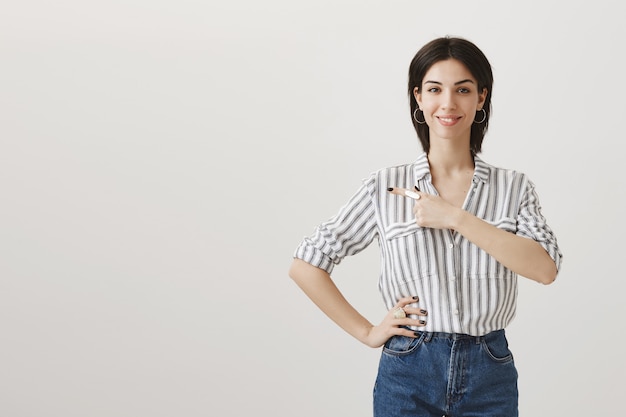 Foto gratuita joven mujer elegante mostrando el anuncio, señalando con el dedo a la izquierda y sonriendo