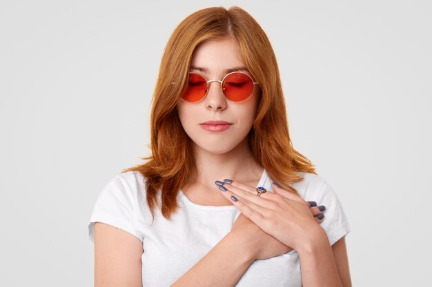 Joven mujer con elegantes gafas de sol, mantiene los ojos cerrados y las manos sobre el corazón, tocada por las buenas palabras dirigidas a ella